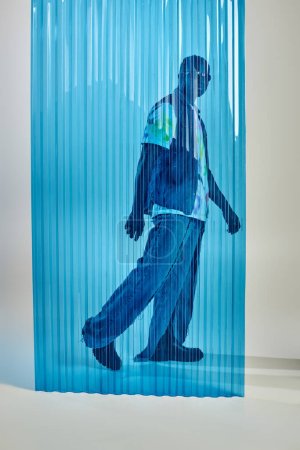 Homme afro-américain élégant en lunettes de soleil, gilet en denim et jean déchiré marchant derrière une feuille de polycarbonate bleu sur fond gris, tournage de mode, vêtements DIY, mode de vie durable 