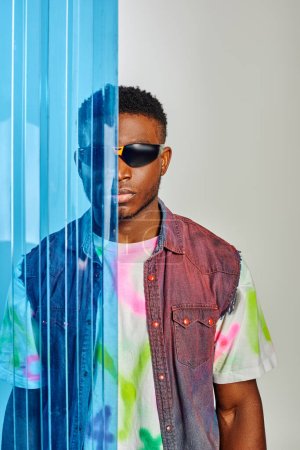Portrait of fashionable young afroamerican man in sunglasses, denim vest and t-shirt standing behind blue polycarbonate sheet on grey background, fashion shoot, DIY clothing, sustainable lifestyle 