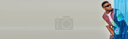 Foto de Modelo afroamericano joven y elegante en gafas de sol, chaleco de mezclilla y camiseta colorida posando detrás de la hoja de policarbonato azul sobre fondo gris, sesión de moda con espacio para copiar, pancarta - Imagen libre de derechos
