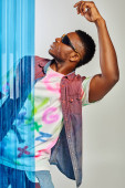 Side view of trendy young afroamerican man in sunglasses, colorful denim vest and t-shirt posing and standing behind blue polycarbonate sheet on grey background, DIY clothing, sustainable lifestyle  Stickers #658610606