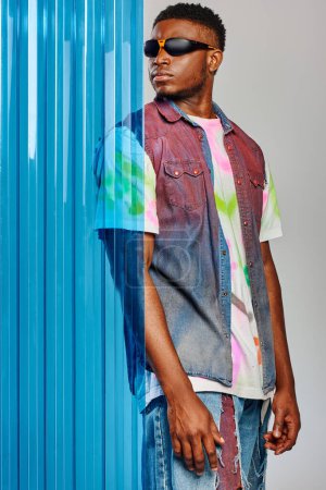 Hombre afroamericano confiado en gafas de sol, chaleco denim colorido y camiseta de pie y posando detrás de la hoja de policarbonato azul sobre fondo gris, sesión de moda, ropa de bricolaje, estilo de vida sostenible 