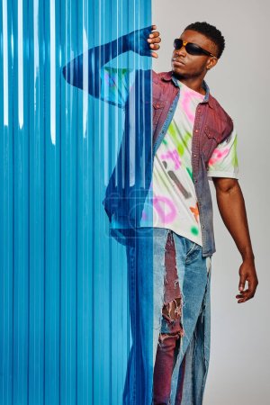 Gut aussehendes junges afroamerikanisches Model in Sonnenbrille, Jeansweste und T-Shirt, das blaues Polycarbonat-Laken berührt und auf grauem Hintergrund steht, Mode-Shooting, DIY-Kleidung, nachhaltiger Lebensstil 