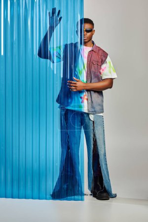 Afroamerikaner mit Sonnenbrille, zerrissenen Jeans und bunter Jeansweste posiert neben blauem Polycarbonat-Laken und steht auf grauem Hintergrund, Mode-Shooting, DIY-Kleidung, nachhaltiger Lebensstil 