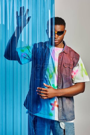 Foto de Modelo masculino afroamericano de moda en gafas de sol, camiseta colorida y chaleco denim posando detrás de la hoja de policarbonato azul sobre fondo gris, sesión de moda, ropa de bricolaje, estilo de vida sostenible - Imagen libre de derechos
