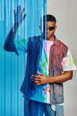 Trendy afroamerican male model in sunglasses, colorful t-shirt and denim vest posing behind blue polycarbonate sheet on grey background, fashion shoot, DIY clothing, sustainable lifestyle  magic mug #658610706