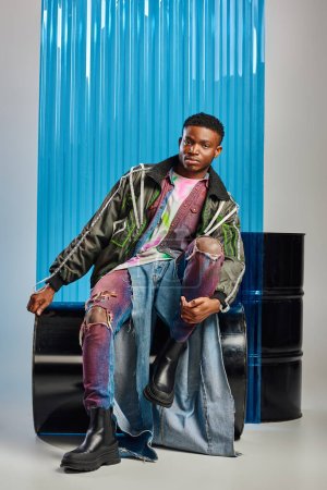Longitud completa de modelo afroamericano joven y elegante en chaqueta de outwear con rayas led y jeans rasgados mirando a la cámara mientras está sentado cerca de la hoja de policarbonato azul en gris, ropa de bricolaje 