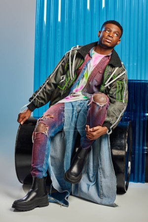 Good looking young afroamerican model in outwear jacket with led stripes and ripped jeans looking away while sitting on fuel barrel near blue polycarbonate sheet on grey background, DIY clothing 