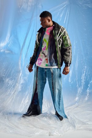 Volle Länge des stylischen afroamerikanischen männlichen Modells in Unterhosen-Jacke mit Bleistreifen und zerrissenen Jeans, die wegschauen, während sie auf glänzendem Zellophan auf blauem Hintergrund stehen, urbanes Outfit, DIY-Kleidung 