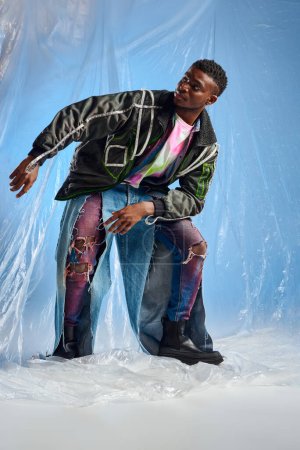 Elegante modelo afroamericano joven en chaqueta de outwear con rayas led y jeans rasgados mirando hacia otro lado y posando sobre celofán brillante sobre fondo azul, atuendo urbano y estilo de vida sostenible 