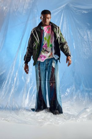 Volle Länge des modischen jungen afroamerikanischen Mannes in Unterhosen-Jacke mit Bleistreifen und zerrissenen Jeans, der in die Kamera schaut, während er auf glänzendem Zellophan auf blauem Hintergrund steht, nachhaltiger Lebensstil 