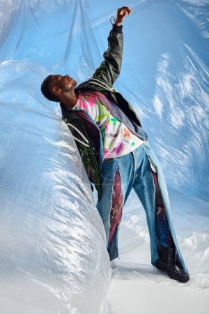 Téléchargez les photos : Vue latérale du beau modèle afro-américain masculin en veste de outwear avec des rayures led et un jean déchiré debout sur un cellophane brillant sur fond bleu, vêtements DIY, mode de vie durable - en image libre de droit
