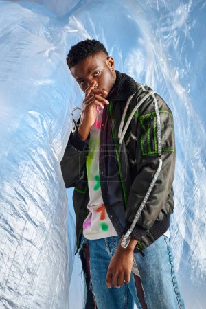 Photo for Confident young afroamerican model looking at camera while covering face and posing in outwear jacket with led stripes and ripped jeans glossy cellophane on blue background, sustainable lifestyle - Royalty Free Image