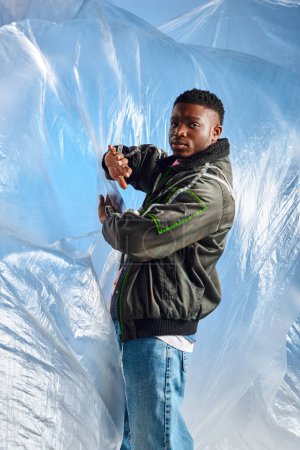 Selbstbewusster junger afroamerikanischer Mann in zerrissenen Jeans und Outwear-Jacke mit LED-Streifen, die glänzendes Zellophan auf blauem Hintergrund berührt, urbanes Outfit und moderne Pose, kreativer Ausdruck, DIY-Kleidung 