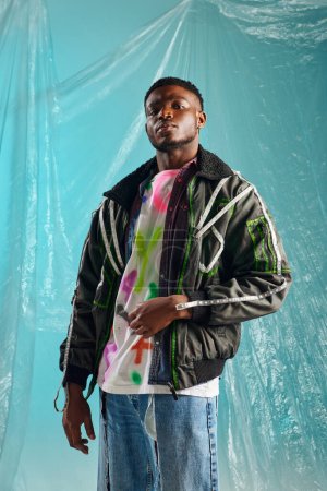 Confident young afroamerican model in ripped jeans and outwear jacket with led stripes looking at camera on glossy cellophane on turquoise background, urban outfit, creative expression, DIY clothing 