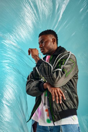 Fashionable young afroamerican model in outwear jacket with led stripes looking away near glossy cellophane on turquoise background, urban outfit and modern pose, creative expression, DIY clothing 