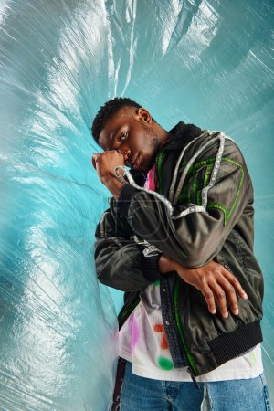 Confident young african american model in outwear jacket with led stripes and ripped jeans posing near glossy cellophane on turquoise background, urban outfit, creative expression, DIY clothing 
