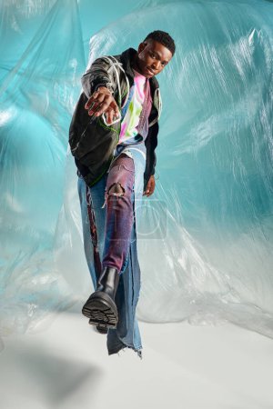 Voller Länge trendiges junges afroamerikanisches Model in Outwear-Jacke und zerrissenen Jeans, das in die Kamera schaut und in der Nähe von glänzendem Zellophan auf türkisfarbenem Hintergrund spaziert, kreativer Ausdruck, DIY-Kleidung 
