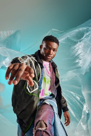 Porträt eines selbstbewussten jungen afroamerikanischen Mannes in Outwear-Jacke und zerrissenen Jeans, der mit Zellophan auf türkisfarbenem Hintergrund posiert, urbanes Outfit und moderne Pose, kreativer Ausdruck, DIY-Kleidung 