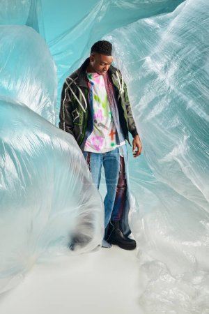 Volle Länge des modischen afroamerikanischen männlichen Modells in Outwear-Jacke mit Bleistreifen und zerrissenen Jeans, die in der Nähe von Zellophan auf türkisfarbenem Hintergrund steht, kreativer Ausdruck, DIY-Kleidung 