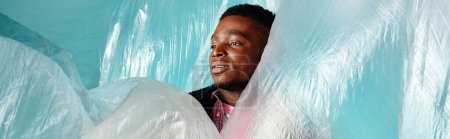 Young african american male model looking away while standing near cellophane on turquoise background, urban outfit and modern pose, banner, creative expression 