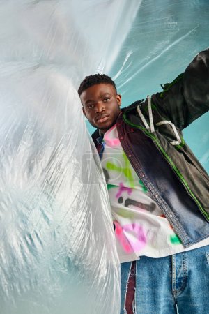 Gut aussehender afroamerikanischer Mann in Outwear-Jacke mit LED-Streifen und buntem T-Shirt und Blick in die Kamera in der Nähe von glänzendem Zellophan auf türkisfarbenem Hintergrund, kreativer Ausdruck, DIY-Kleidung 