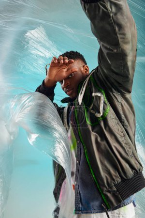 Stylish afroamerican model in outwear jacket with led stripes covering face while looking at camera near cellophane on turquoise background, urban outfit, creative expression, DIY clothing 