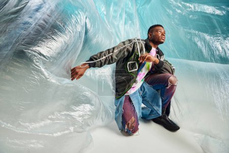 Jeune modèle afro-américain en veste de outwear à rayures led et jean déchiré regardant loin près de cellophane sur fond turquoise, tenue urbaine et pose moderne, expression créative, vêtements DIY 