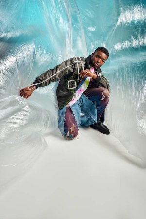 Modelo afroamericano joven de moda y confiado en chaqueta de outwear y pantalones vaqueros rasgados mirando la cámara cerca de celofán sobre fondo turquesa, expresión creativa, ropa de bricolaje 