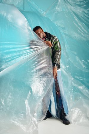 Photo for African american man in ripped jeans and outwear jacket with led stripes covering face with cellophane on turquoise background, urban outfit and modern pose, creative expression, DIY clothing - Royalty Free Image
