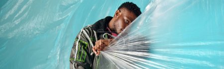 Young and trendy afroamerican model in outwear jacket and led stripes covering face with cellophane on turquoise background, urban outfit and modern pose, banner, creative expression, DIY clothing 