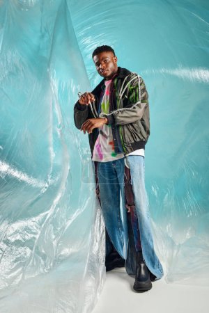 Full length of stylish afroamerican man in ripped jeans touching sleeve of outwear jacket with led stripes near cellophane on turquoise background, urban outfit and modern pose, DIY clothing 