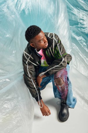 Homme afro-américain à la mode en veste de outwear à rayures led et jeans déchirés à la mode regardant loin près de cellophane brillant sur fond turquoise, tenue urbaine et pose moderne, expression créative