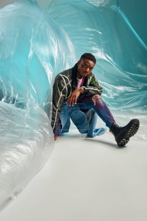 Foto de Joven modelo afroamericano en chaqueta de outwear con rayas led y jeans rasgados sentados cerca de celofán brillante sobre fondo turquesa, atuendo urbano y pose moderna, expresión creativa, ropa de bricolaje - Imagen libre de derechos