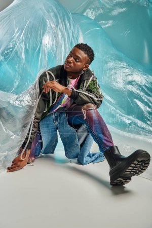 Modèle masculin afro-américain à la mode en jean déchiré et veste de sport avec rayures led détournant les yeux et posant près de cellophane sur fond turquoise, expression créative, vêtements de bricolage 