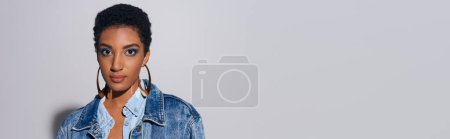 Foto de Retrato de modelo afroamericano joven de moda con maquillaje audaz en chaqueta de mezclilla y pendientes de oro mirando a la cámara en fondo gris con espacio de copia, concepto de moda de mezclilla, pancarta - Imagen libre de derechos