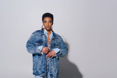 Fashionable african american woman with short hair looking at camera while posing in denim jacket and golden earrings on grey background, denim fashion concept