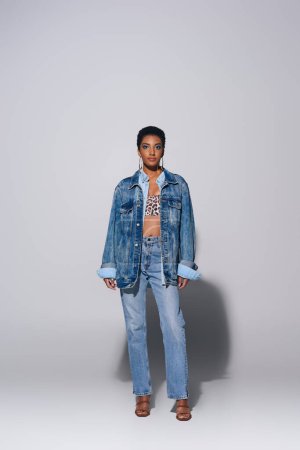 Longitud completa de la mujer afroamericana de moda con pelo corto y maquillaje audaz posando en chaqueta de mezclilla y jeans mientras está de pie sobre fondo gris, concepto de moda de mezclilla