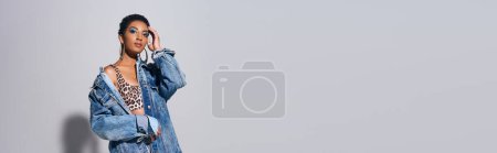 Téléchargez les photos : Jeune femme afro-américaine aux cheveux courts avec un maquillage audacieux et des boucles d'oreilles dorées touchant la tête tout en posant en haut et veste en denim sur fond gris, concept de mode en denim, bannière - en image libre de droit
