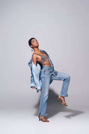 Full length of fashionable african american woman with bold makeup posing in top with animal print, denim jacket and jeans while standing on grey background, denim fashion concept