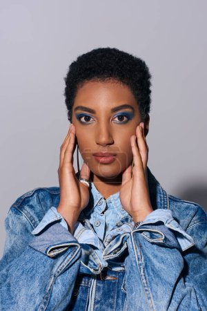 Téléchargez les photos : Jeune modèle afro-américain au maquillage vif et aux cheveux courts touchant les joues et posant en denim tout en se tenant debout sur fond gris, concept de mode denim - en image libre de droit