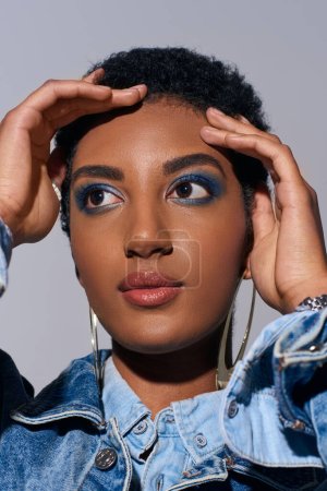 Portrait of stylish african american model with bold makeup and golden earrings touching short hair and looking away while posing in denim outfit isolated on grey, denim fashion concept