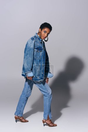 Longitud completa de la mujer afroamericana joven de moda con pendientes de oro mirando a la cámara mientras posa en jeans y chaqueta de mezclilla sobre fondo gris, concepto de moda de mezclilla