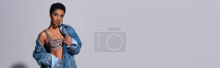 Foto de Moderna mujer afroamericana de pelo corto en la parte superior con estampado de animales y ropa de mezclilla mirando a la cámara y de pie sobre fondo gris, concepto de moda de mezclilla, bandera - Imagen libre de derechos