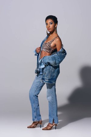 Full length of fashionable african american woman with bold makeup wearing top with animal print, denim jacket and jeans on grey background, denim fashion concept