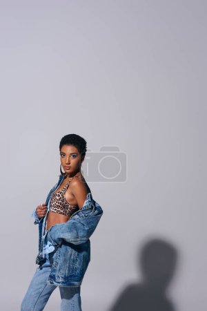 Fashionable african american woman with golden earrings and short hair posing in top with animal print and denim jacket on grey background, denim fashion concept