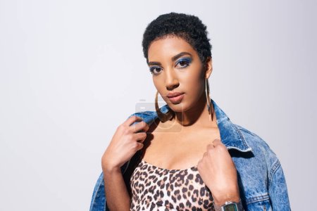 Young and short haired african american woman with blue eyeshadow and golden earrings posing in top with animal print and wearing denim jacket isolated on grey, denim fashion concept