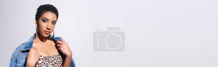 Foto de Mujer afroamericana de moda con maquillaje vívido y pendientes dorados que usan chaqueta de mezclilla mientras posan en la parte superior con estampado animal aislado en gris, concepto de moda de mezclilla, pancarta - Imagen libre de derechos