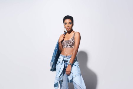 Confident young african american model with short hair and vivid makeup posing in top with leopard print and denim outfit while standing on grey background, denim fashion concept