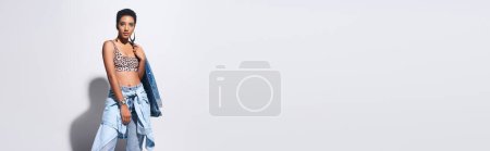 Foto de Modelo afroamericano joven de moda con pelo corto posando en la parte superior con impresión animal y ropa de mezclilla sobre fondo gris con espacio de copia, concepto de moda de mezclilla, pancarta - Imagen libre de derechos