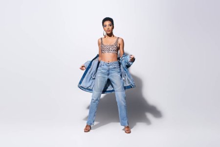 Full length of trendy and short haired african american model in top with leopard print and jeans posing with denim jacket while standing on grey background, denim fashion concept